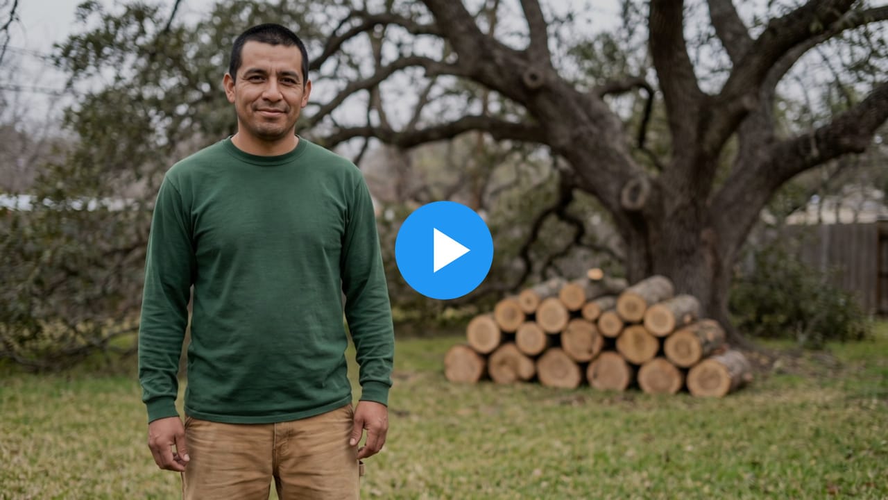 Miguel, a tree service owner, in a back yard in front of an oak tree and a stack of cut logs